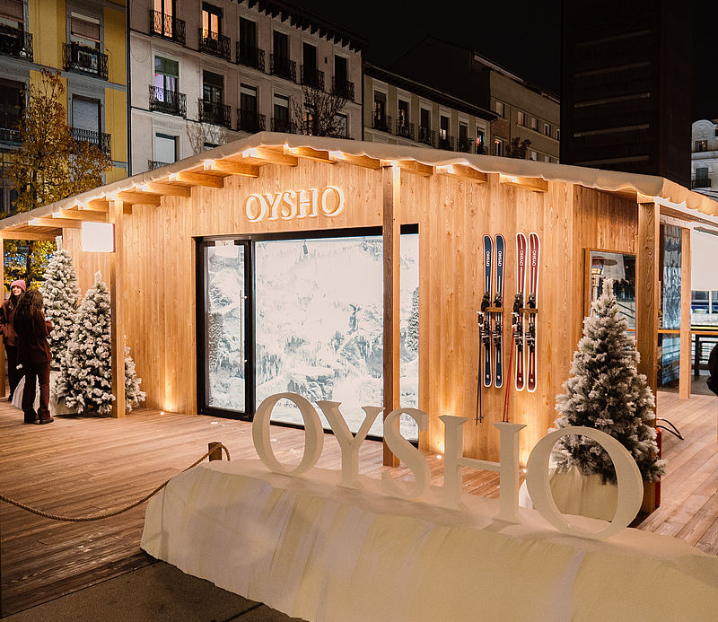 Beleuchtete Oysho-Holzhütte bei Nacht mit OYSHO-Schriftzug, Schnee und Tannenbäumen in Madrid
