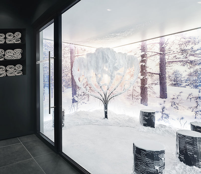 Blick durch Glasfront in Schneeraum mit verschneitem Baum, Holzhockern und Handtuchregal.