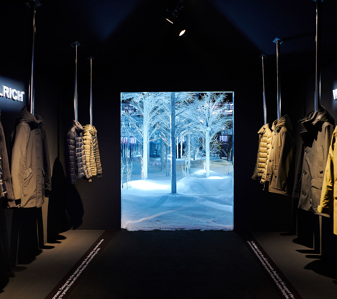 Boutique Woolrich avec vestes sur portants et image forêt hivernale illuminée en arrière-plan