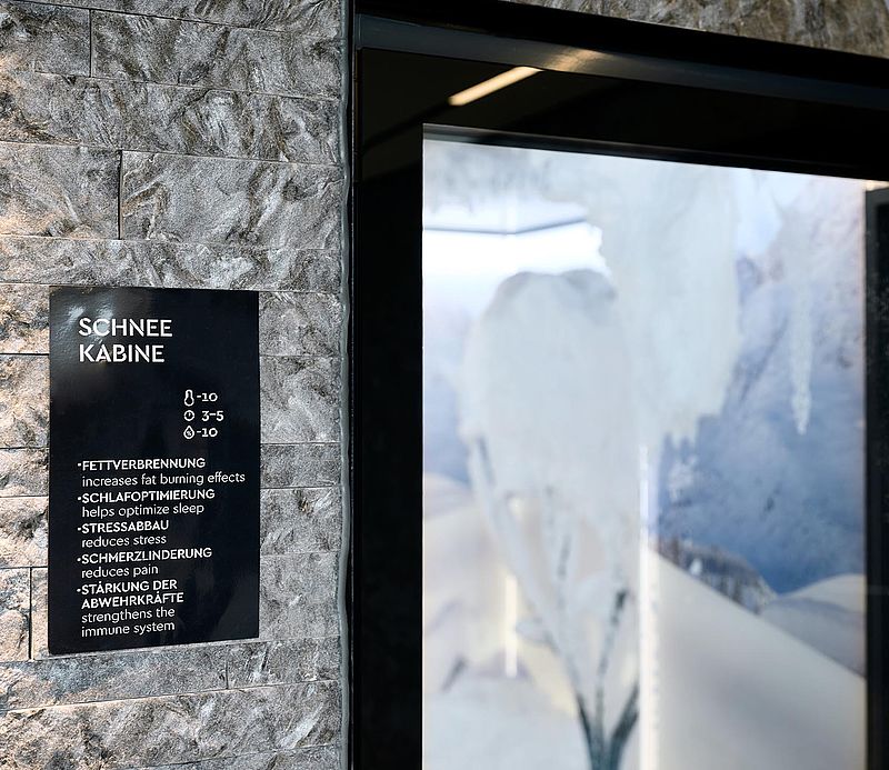 Eingangsschild Schneekabine an Steinwand mit Glastür und Blick auf verschneiten Innenraum.