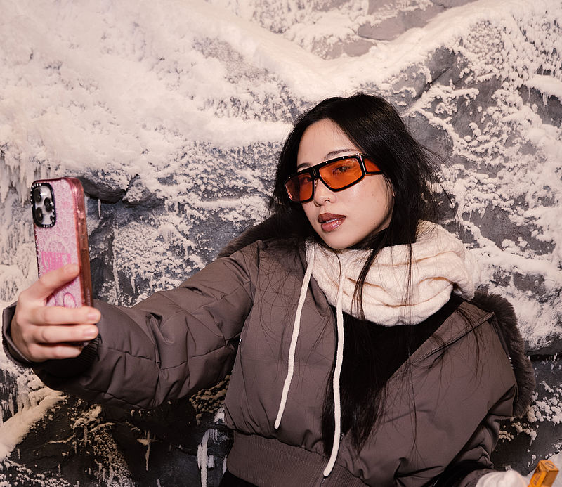 Junge Frau mit Sonnenbrille macht Selfie vor verschneiter Eiswand im Oysho Coldroom