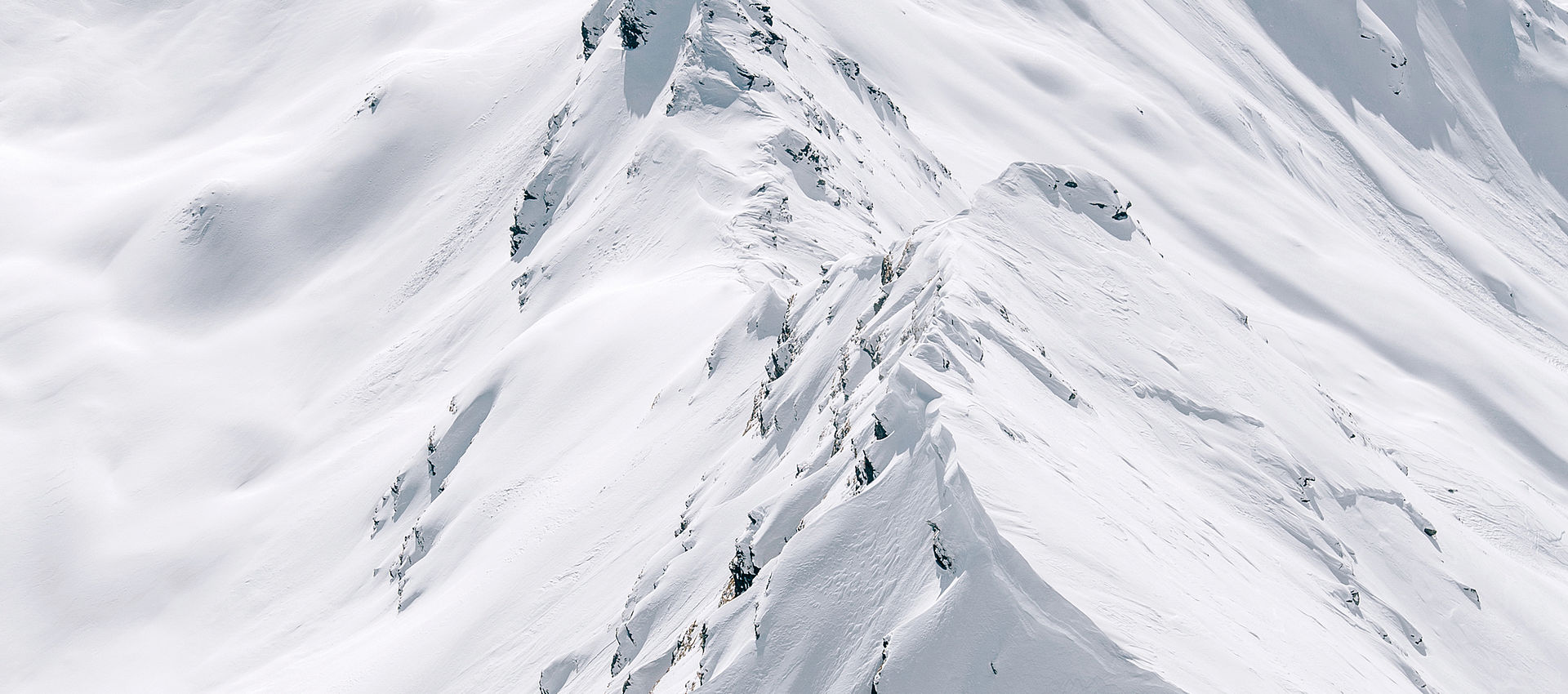 Aerial view of dramatic snow-covered alpine mountain ridges and peaks with sharp ridgelines and deep snow blanketing rugged terrain under pale sky