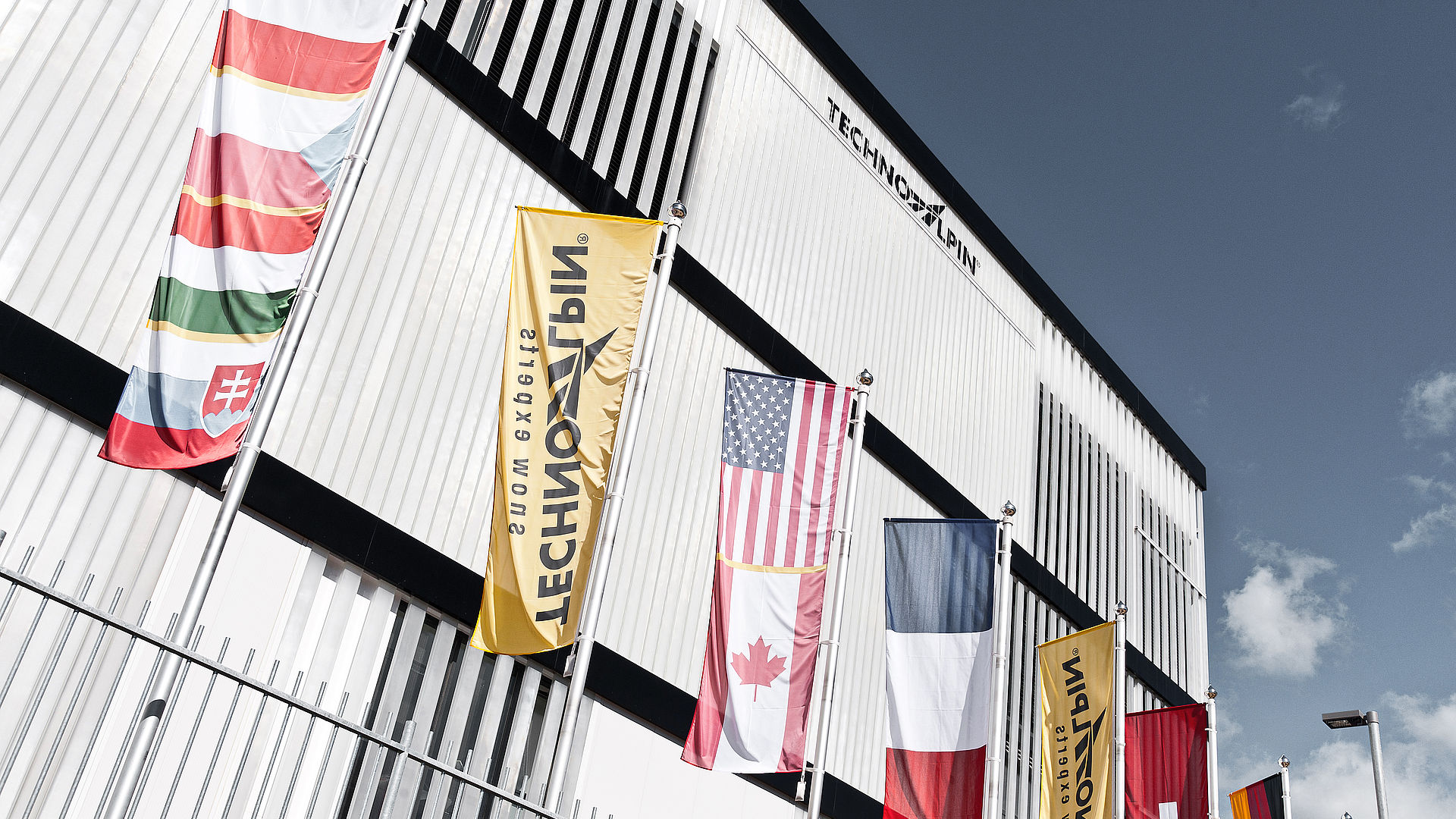 TechnoAlpin Firmengebäude mit internationalen Flaggen verschiedener Länder vor blauem Himmel