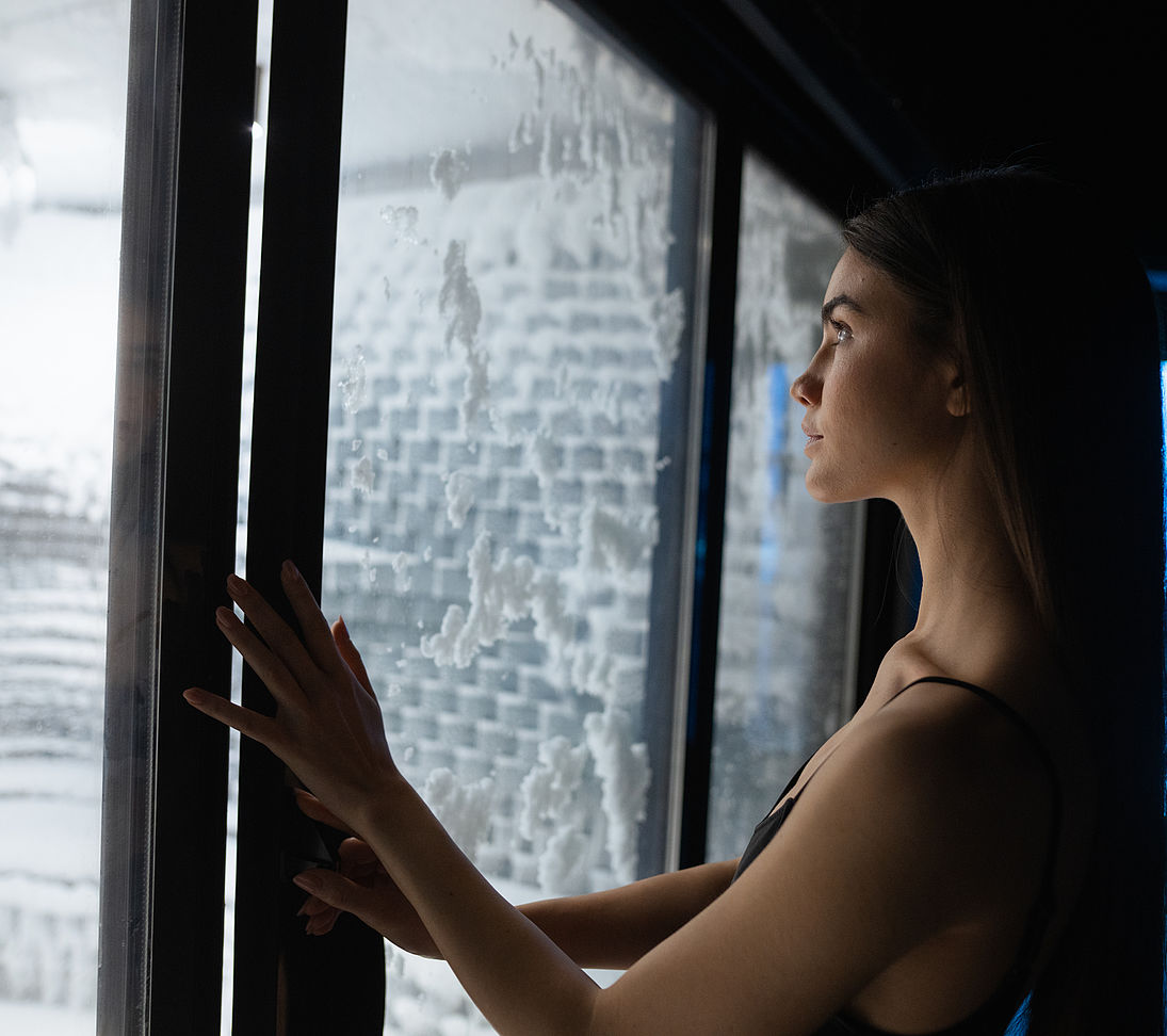 Woman looks through glass door at snowy snow room with white snow wall in spa