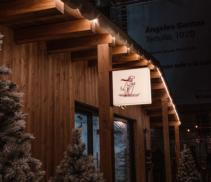 Detail der beleuchteten Oysho-Holzhütte bei Nacht mit verschneiten Tannen und Holzbalkendach