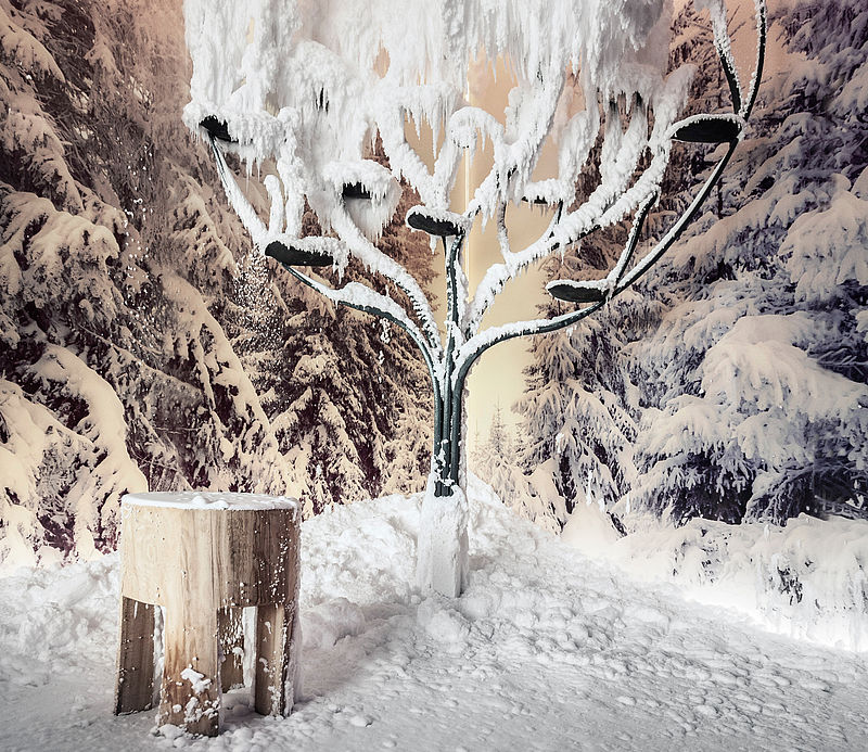 Schneekabine mit kunstvollem vereistem Baum, Holzhocker und Fußspuren im Pulverschnee.