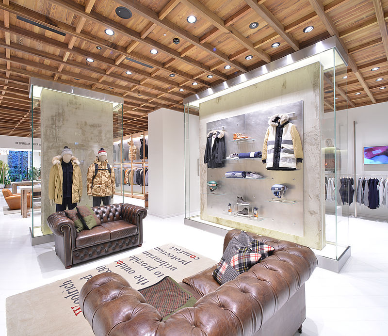 Eleganter Store-Innenraum mit braunen Chesterfield-Sofas, Glasvitrinen und Mannequins unter Holzbalkendecke.