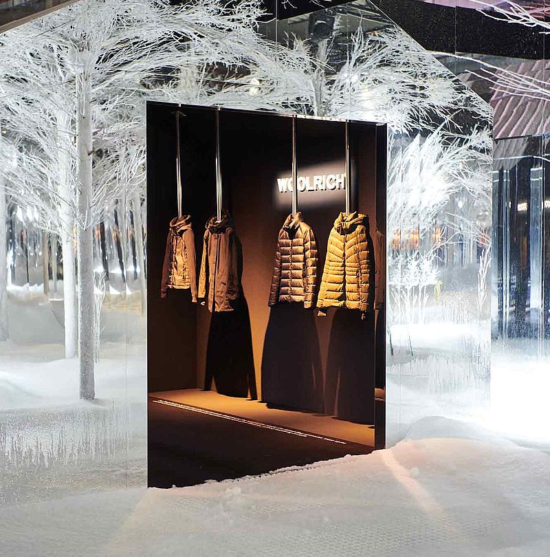 Verschneite Bäume umgeben Woolrich-Schaukasten mit Jacken bei der SnowRoom-Installation
