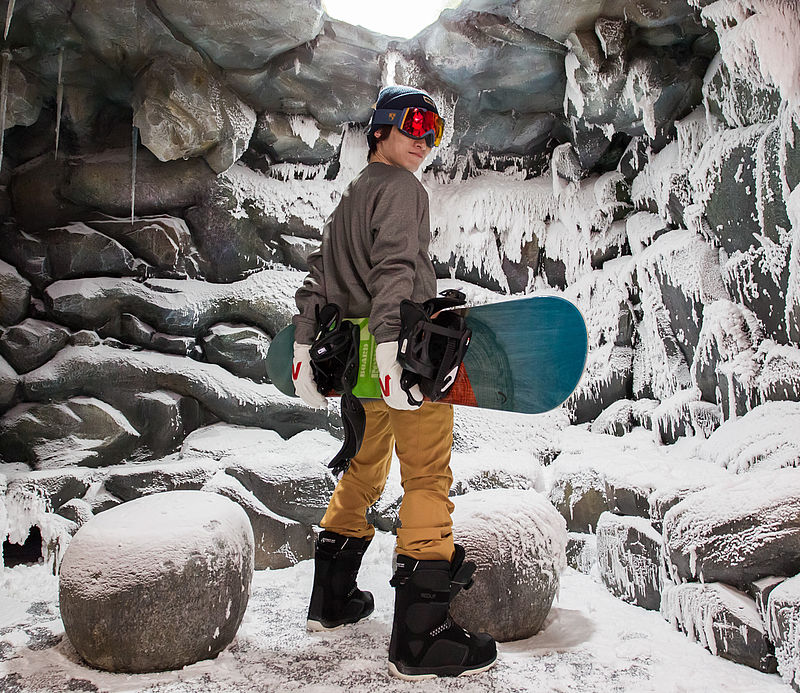 Snowboarder with helmet and goggles in icy indoor snow cave with icicles and snow-covered rocks.